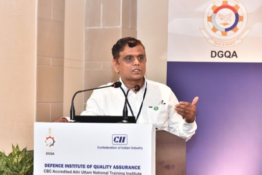 A man in a white shirt speaks at a podium with microphones during a Defence Institute of Quality Assurance event. A banner with the DGQA logo and institutional text is visible in the background.