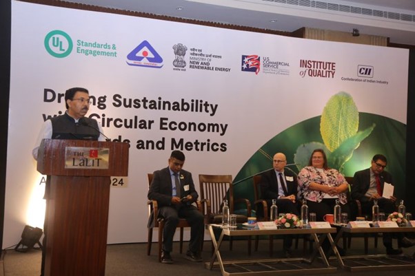 A man speaks at a podium during a conference on sustainability, with four panelists seated on stage and event banners from various organizations displayed behind them.