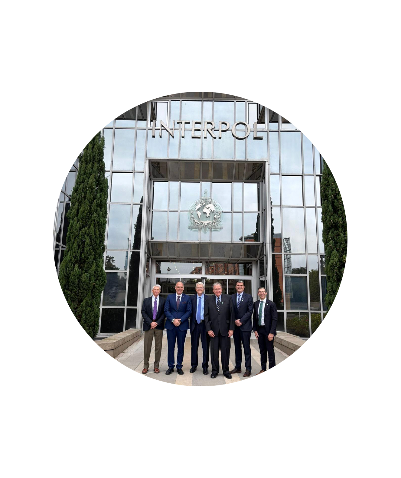Five men in suits stand in front of the INTERPOL headquarters, posing for a group photo. The glass facade and INTERPOL sign are visible above them, highlighting their commitment to anti-counterfeiting and tackling counterfeit risks. Tall trees flank the entrance.