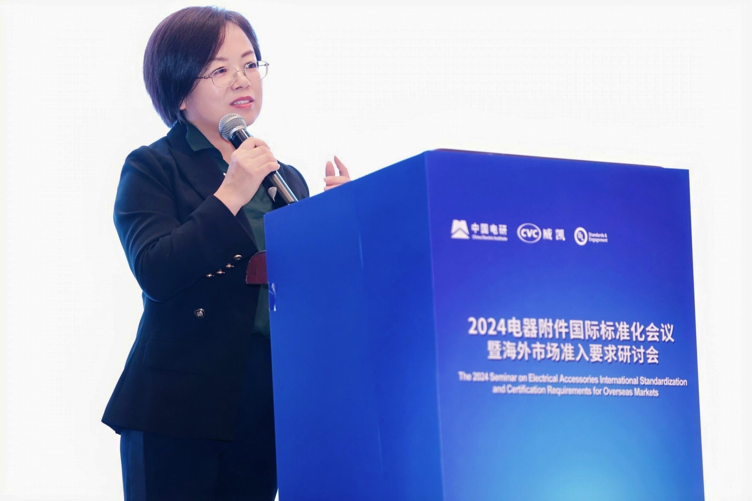 A woman in a black blazer speaks into a microphone at a podium labeled with Chinese text and the English title, "2024 Seminar on Electrical Accessories International Standardization and Certification Requirements for Overseas Markets.