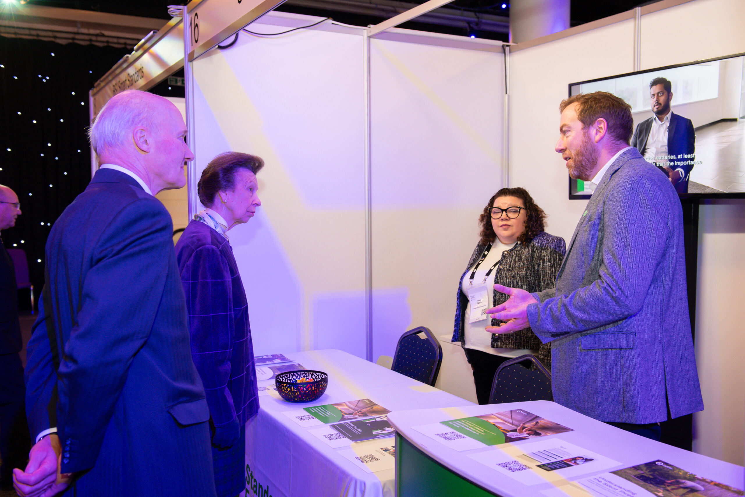 A man and a woman stand behind a booth table talking to two visitors at an exhibition. The table has brochures and a bowl on it, and a TV screen on the wall displays a man speaking.