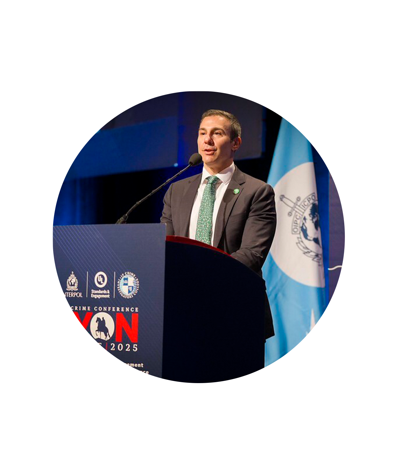 A man in a suit and green tie speaks at a podium with the INTERPOL logo and a blue and white flag, addressing counterfeit risks to intellectual property rights during a crime conference event.