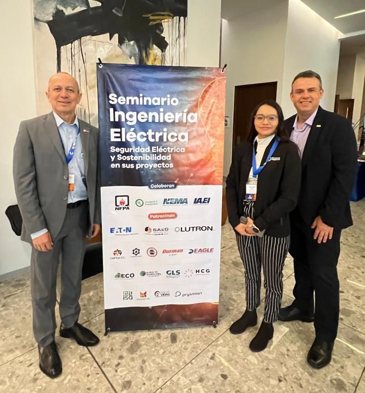 Three people in business attire stand next to a vertical banner for the "Seminario Ingeniería Eléctrica" in a modern, well-lit lobby. The banner displays sponsor logos and seminar details.