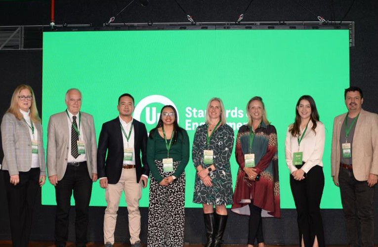 Eight people stand in a row, smiling, in front of a large green screen displaying the words "UI Standards Emprende." They are dressed in business attire and wearing conference badges.