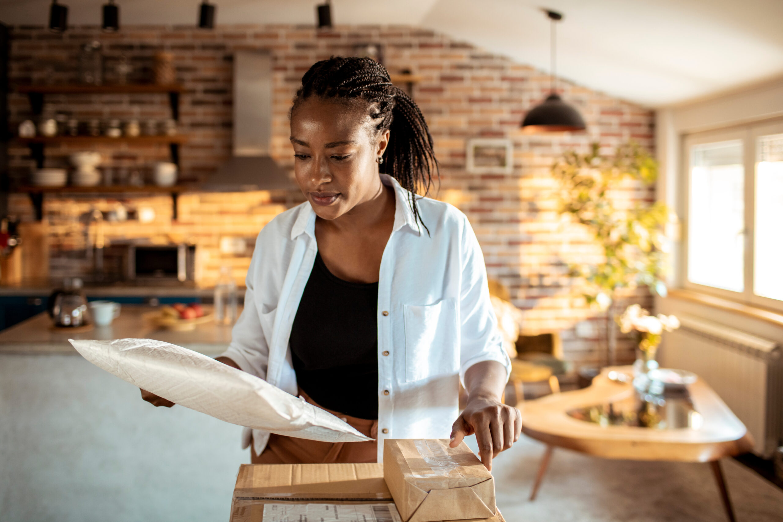 A woman stands in a sunlit kitchen, looking at a padded envelope while holding a cardboard box. She appears to be examining her mail or packages on a wooden table, with shelves and kitchenware visible in the background.