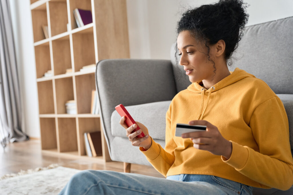 A young woman in a yellow hoodie sits on the floor at home, holding a credit card in one hand and a smartphone in the other, appearing to make an online purchase. A bookshelf and a sofa are visible in the background.