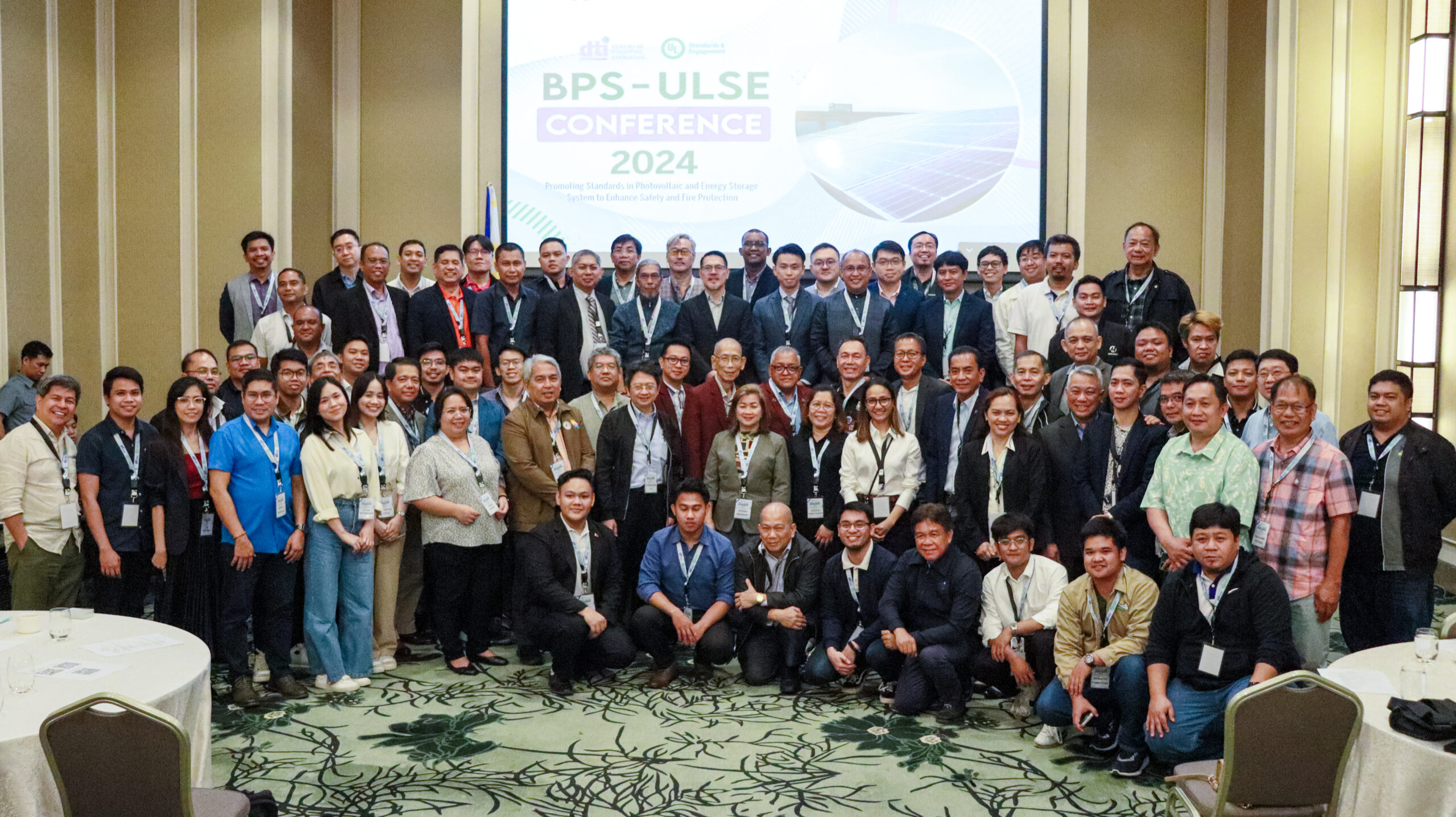 A large group of people poses together in formal and business attire for a group photo at the BPS-ULSE Conference 2024, with a conference banner displayed on a screen behind them in a hotel function room.