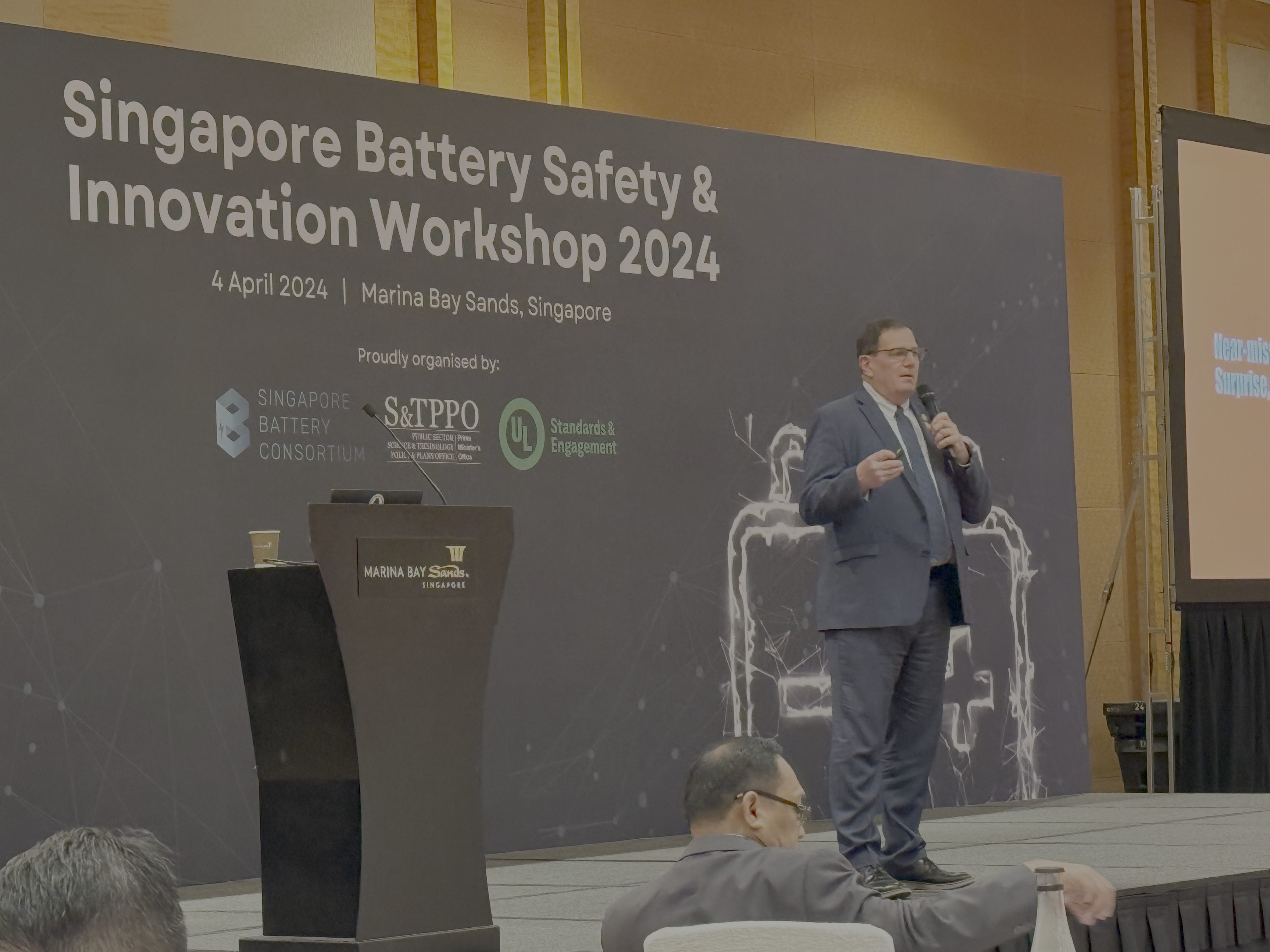 A man in a suit speaks on stage at the Singapore Battery Safety & Innovation Workshop 2024, with a large screen and event banner behind him. Attendees sit in the foreground.