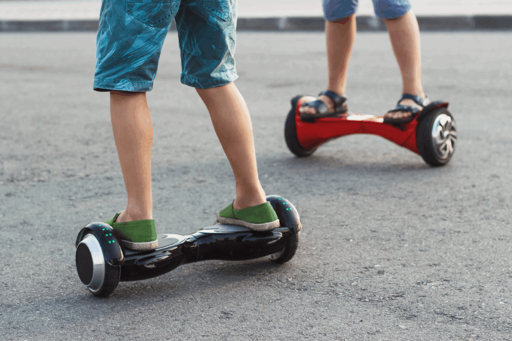 Two people riding hoverboards on a paved surface. One person is on a black hoverboard wearing green shoes and shorts, while the other is on a red hoverboard wearing sandals and blue shorts. Only their legs are visible.