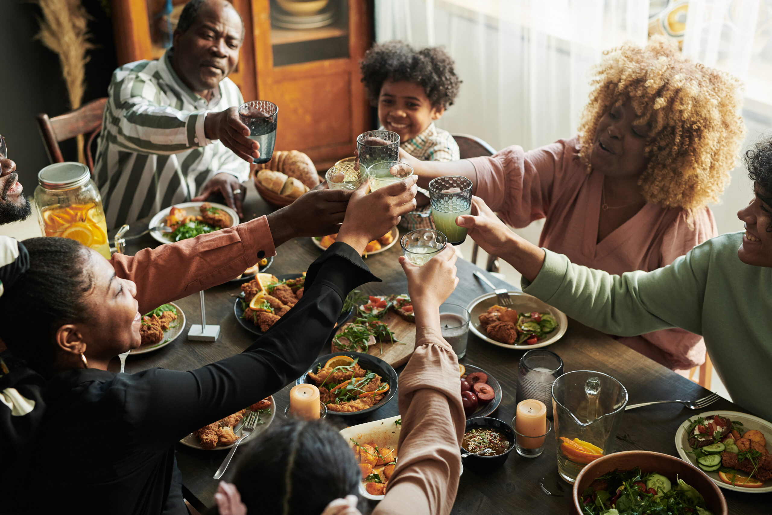 A group of people sitting around a dinner table filled with food, raising their glasses in a celebratory toast, smiling and enjoying each other's company.