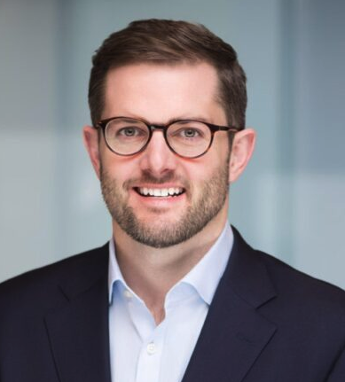 A man with short brown hair, a beard, and glasses is smiling. He is wearing a dark blazer and a light blue shirt. The background is blurred with light blue and white tones.