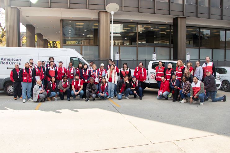 A large group of people wearing red American Red Cross vests gather in a parking lot in front of Red Cross and UL Solutions vehicles.