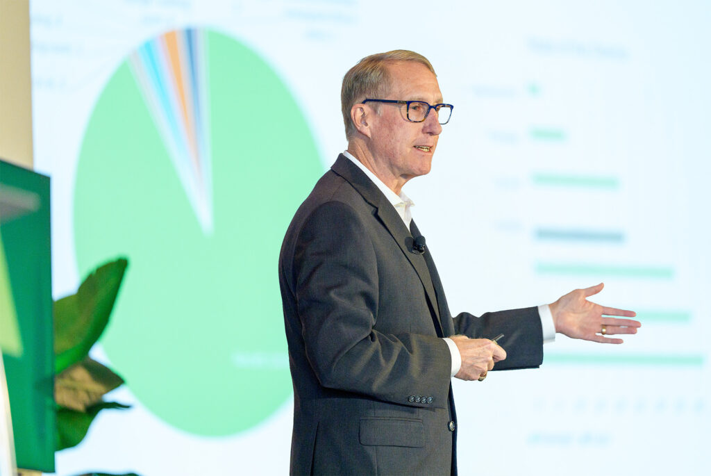A man in a suit and glasses gives a presentation, gesturing toward a screen displaying a large green pie chart and bar graphs as experts discuss insights about UL Standards & Engagement.