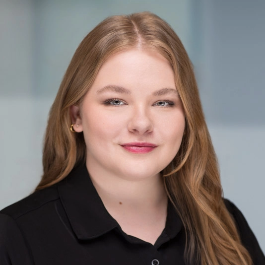 A young woman with long, light brown hair and fair skin, wearing a black collared shirt, poses in front of a blurred, light-colored background. She is smiling softly and looking directly at the camera.