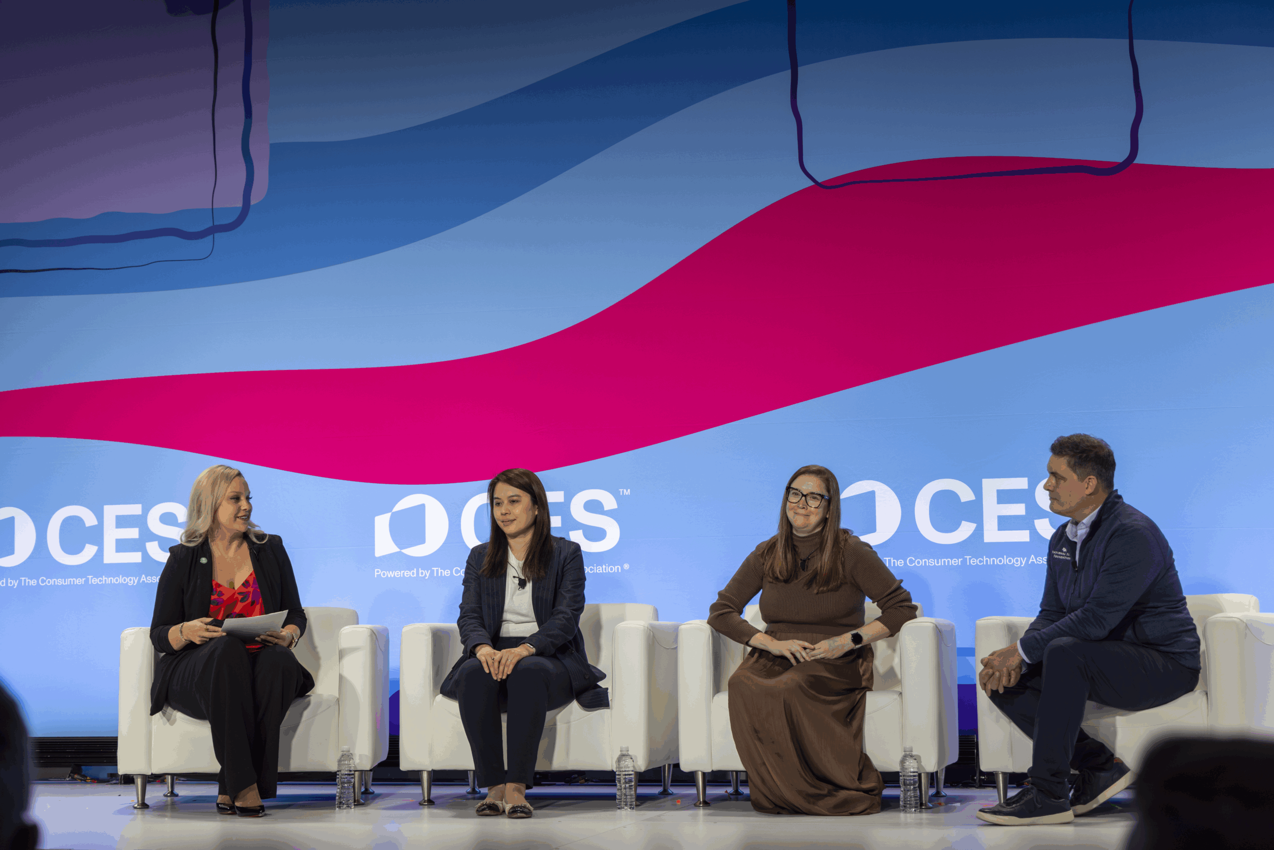 Four people sit on a stage in white chairs, engaged in a panel discussion at CES. Behind them is a colorful, abstract backdrop with the CES logo visible. Three women and one man face the audience, holding notepads and microphones.