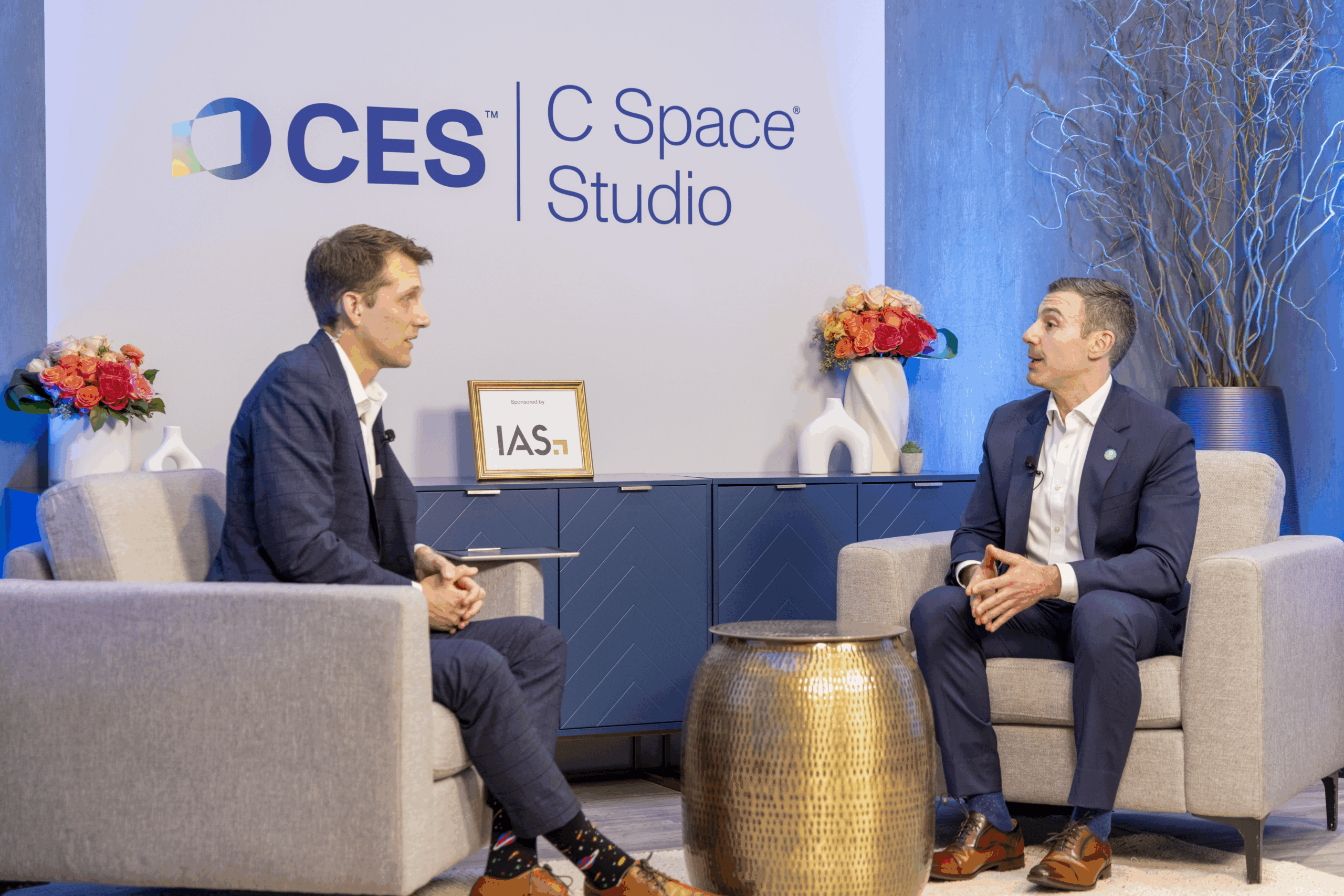 Two men in suits have a conversation while seated in a studio with CES C Space Studio signage behind them, modern decor, flowers, and a gold table between them.