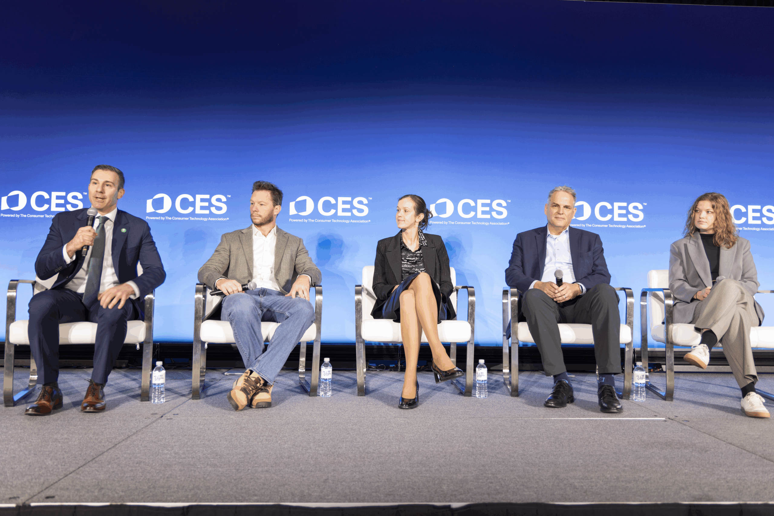 Five people sit on a stage in front of a blue CES backdrop, participating in a panel discussion. One person speaks into a microphone while the others listen. Water bottles are placed beside each chair.