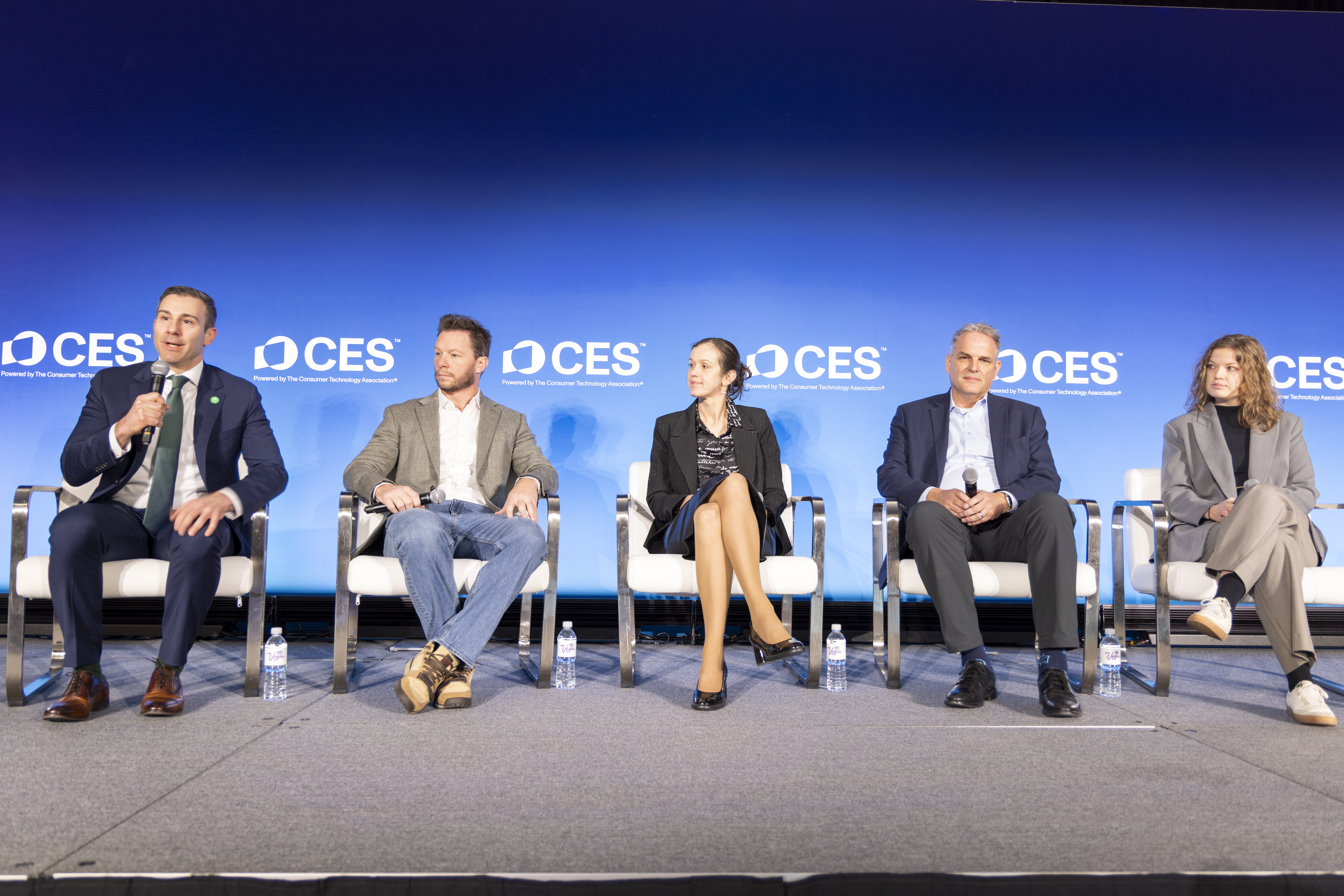 Five people sit on a stage in front of a blue CES backdrop, participating in a panel discussion. One person speaks into a microphone while the others listen. Water bottles are placed beside each chair.