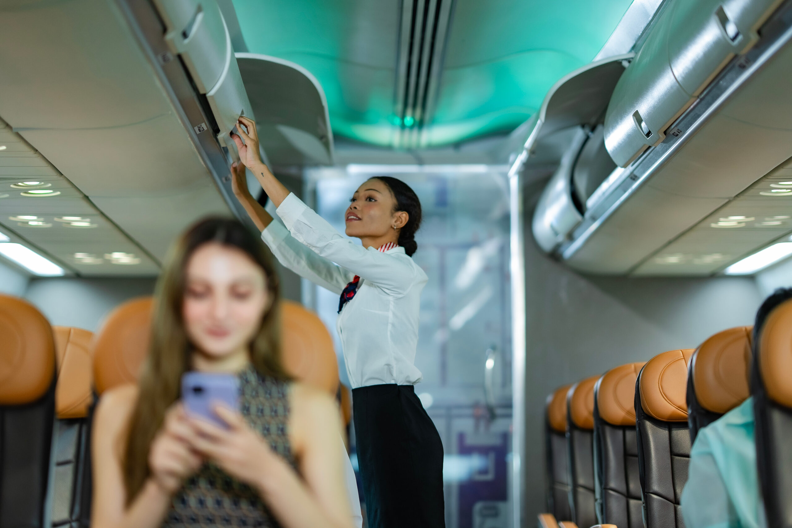 A flight attendant in uniform closes an overhead compartment inside an airplane, while a seated passenger in the foreground uses a smartphone. The cabin has brown leather seats and is well-lit.