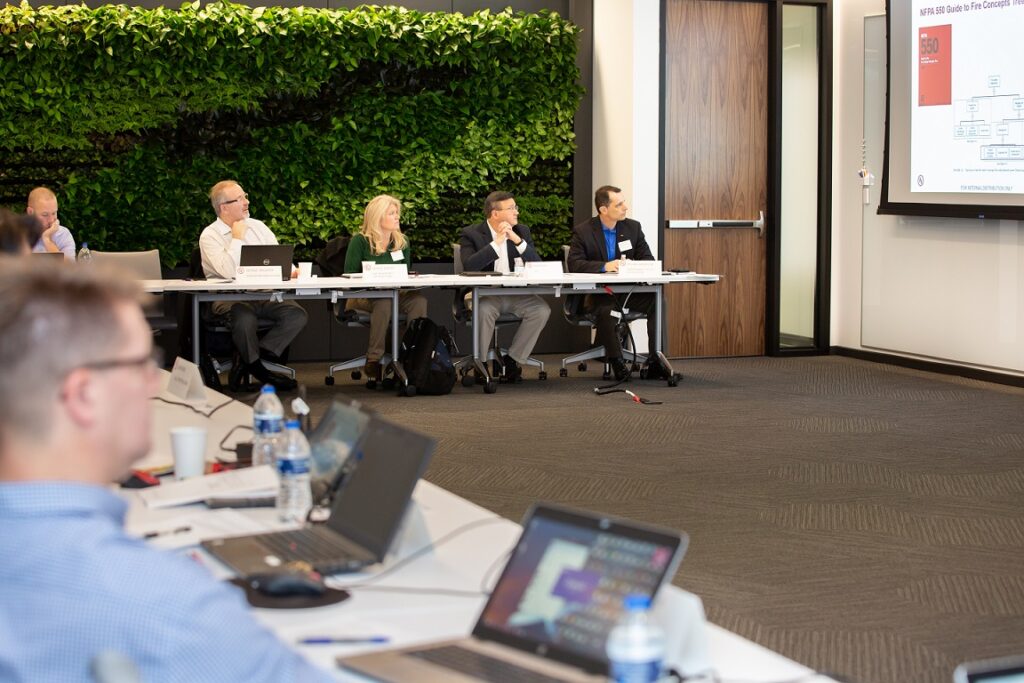 People sitting at a conference table in an office, attentively watching a presentation on UL Standards & Engagement projected on a screen. Laptops, papers, and water bottles are on the tables. A green wall with plants is in the background.
