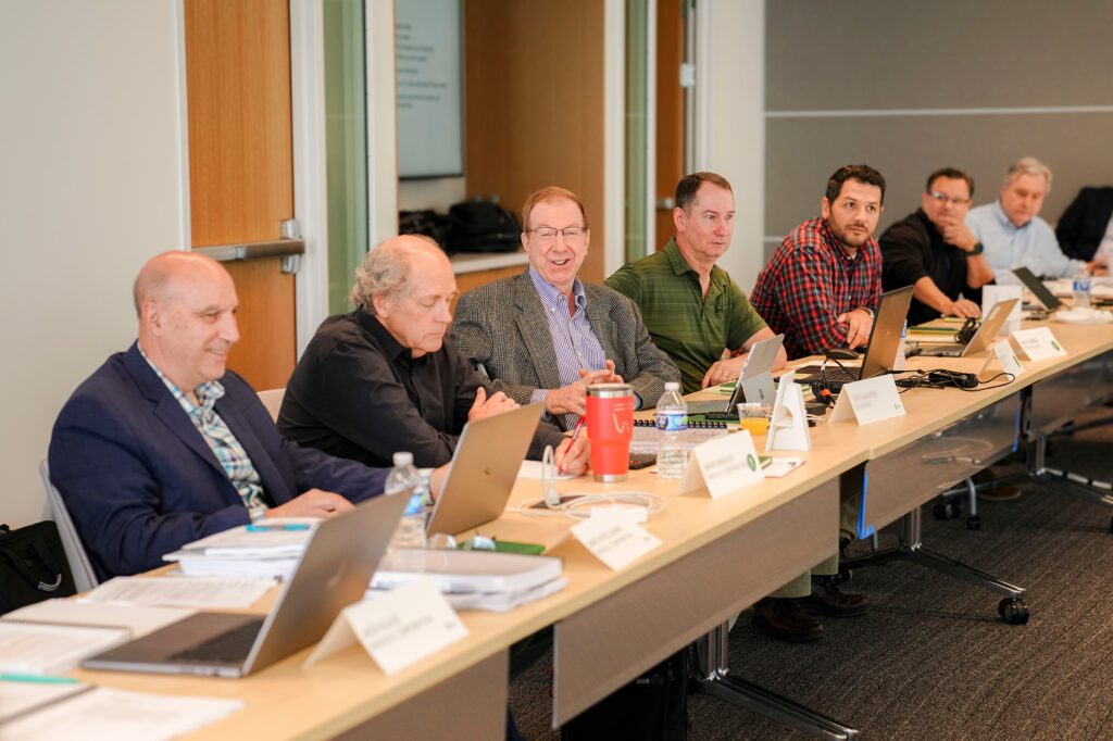 A group of men sit at a conference table with laptops, papers, and name cards, engaged in a meeting. Some are smiling, and the atmosphere appears professional and collaborative.