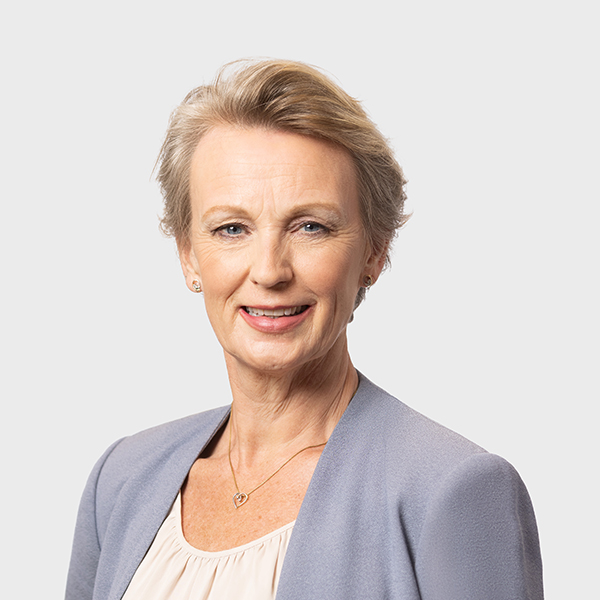 A woman with short, light brown hair smiles at the camera. She is wearing a light purple blazer over a cream blouse and a gold necklace with a heart-shaped pendant, set against a plain light background.