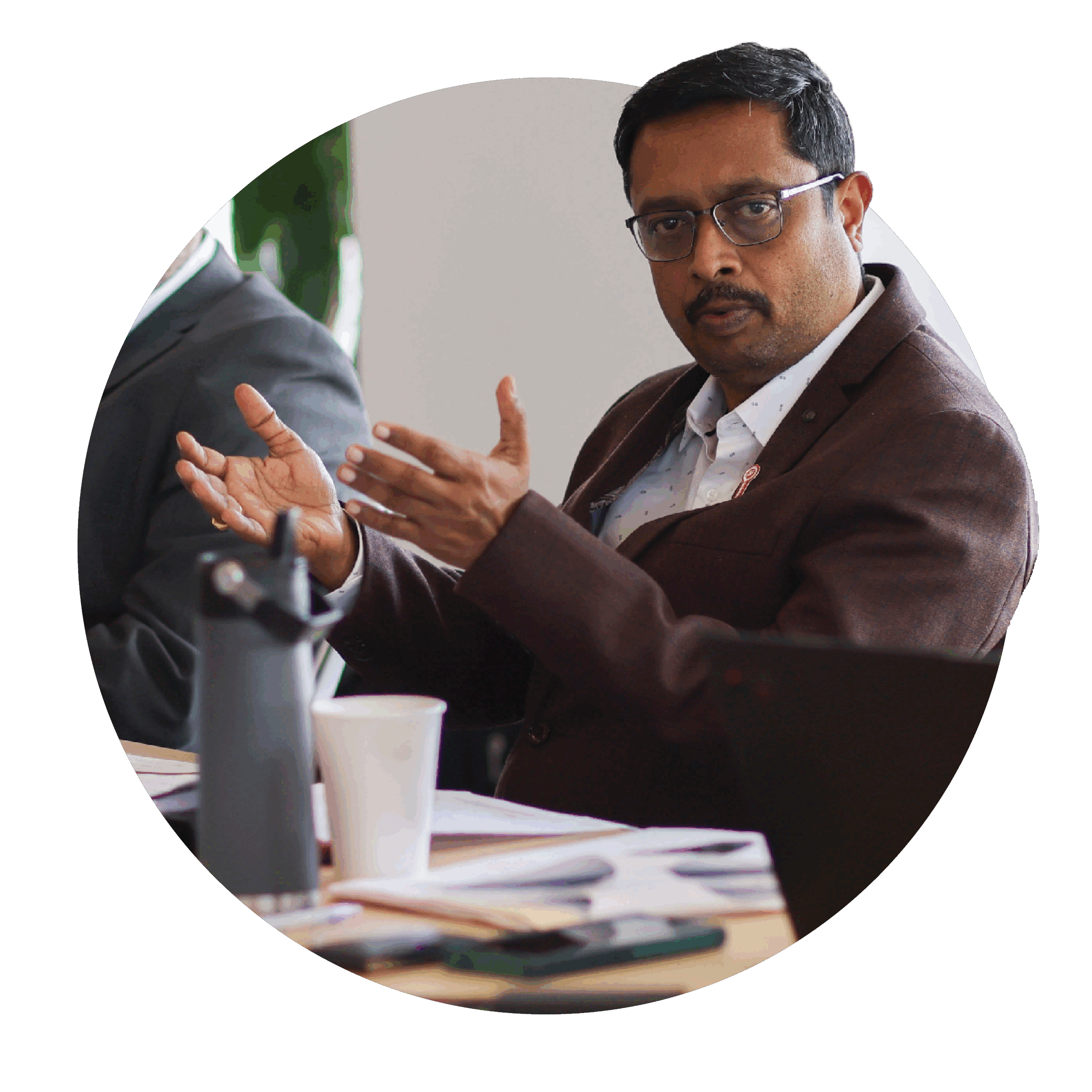 A man in a suit sits at a table, gesturing as he speaks during a UL Standards & Engagement Technical Committee meeting. Papers, a laptop, water bottle, and coffee cup are on the table, highlighting an active standards development discussion.