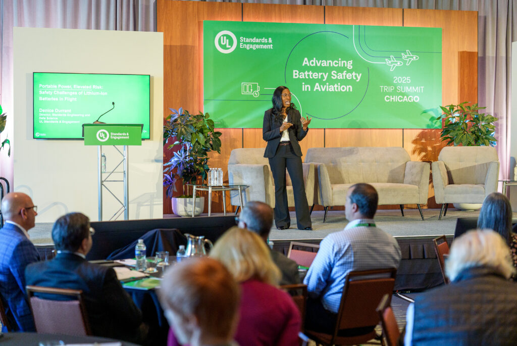 A woman speaks on stage at a conference titled “Advancing Battery Safety in Aviation,” with seated audience members, informational screens, and a banner for the 2025 TRIP Summit in Chicago.