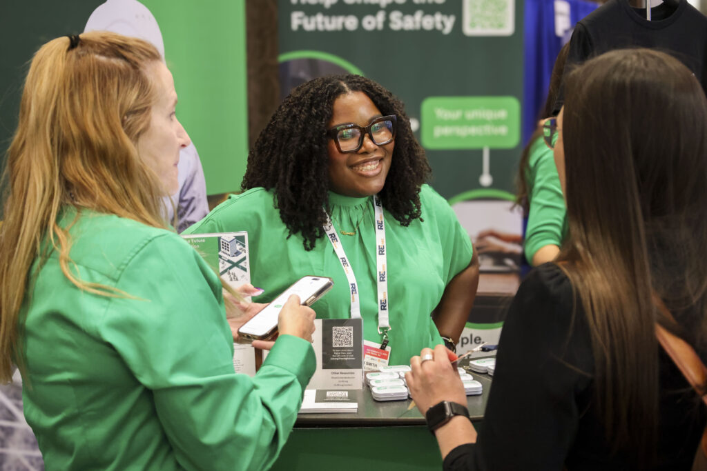 Three women are talking and smiling at a booth, with one woman in green standing behind the counter. The booth displays promotional materials and a green banner highlighting UL Standards & Engagement Culture and Values in the background.