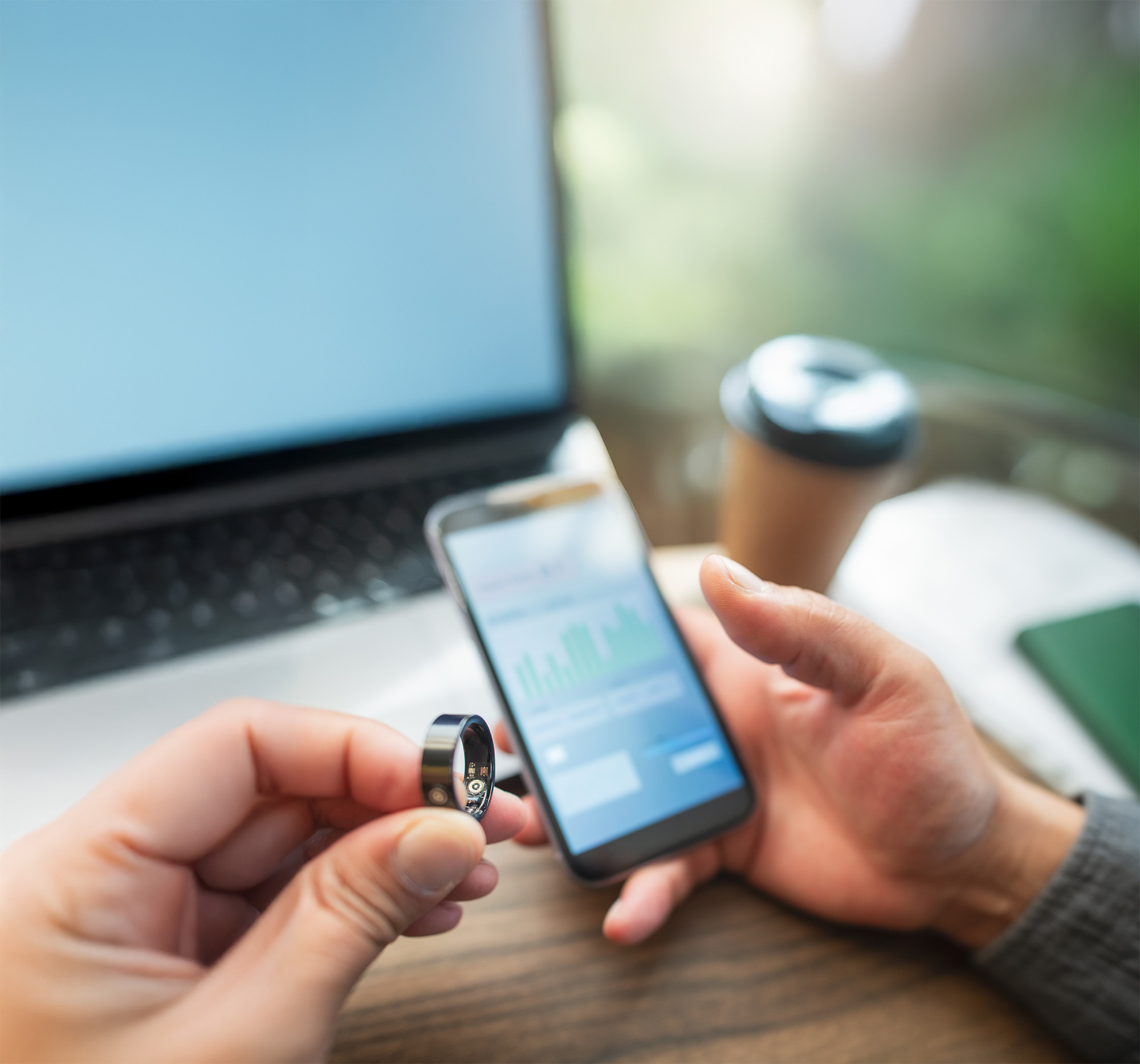 A person holds a ring while another uses a smartphone in front of an open laptop and a coffee cup on a wooden table, suggesting a decision or transaction involving the ring.