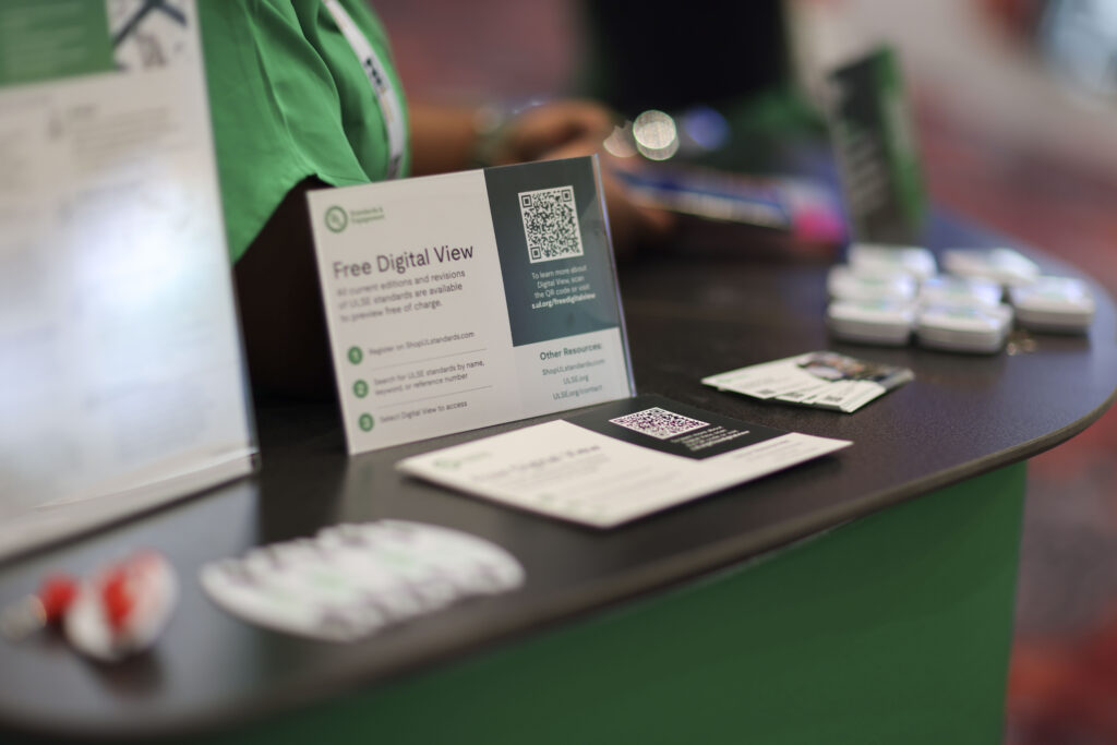 Close-up of a tabletop at an event, displaying brochures, flyers, a QR code sign offering a free digital view, and small promotional items, with a person in a green shirt standing behind the table.