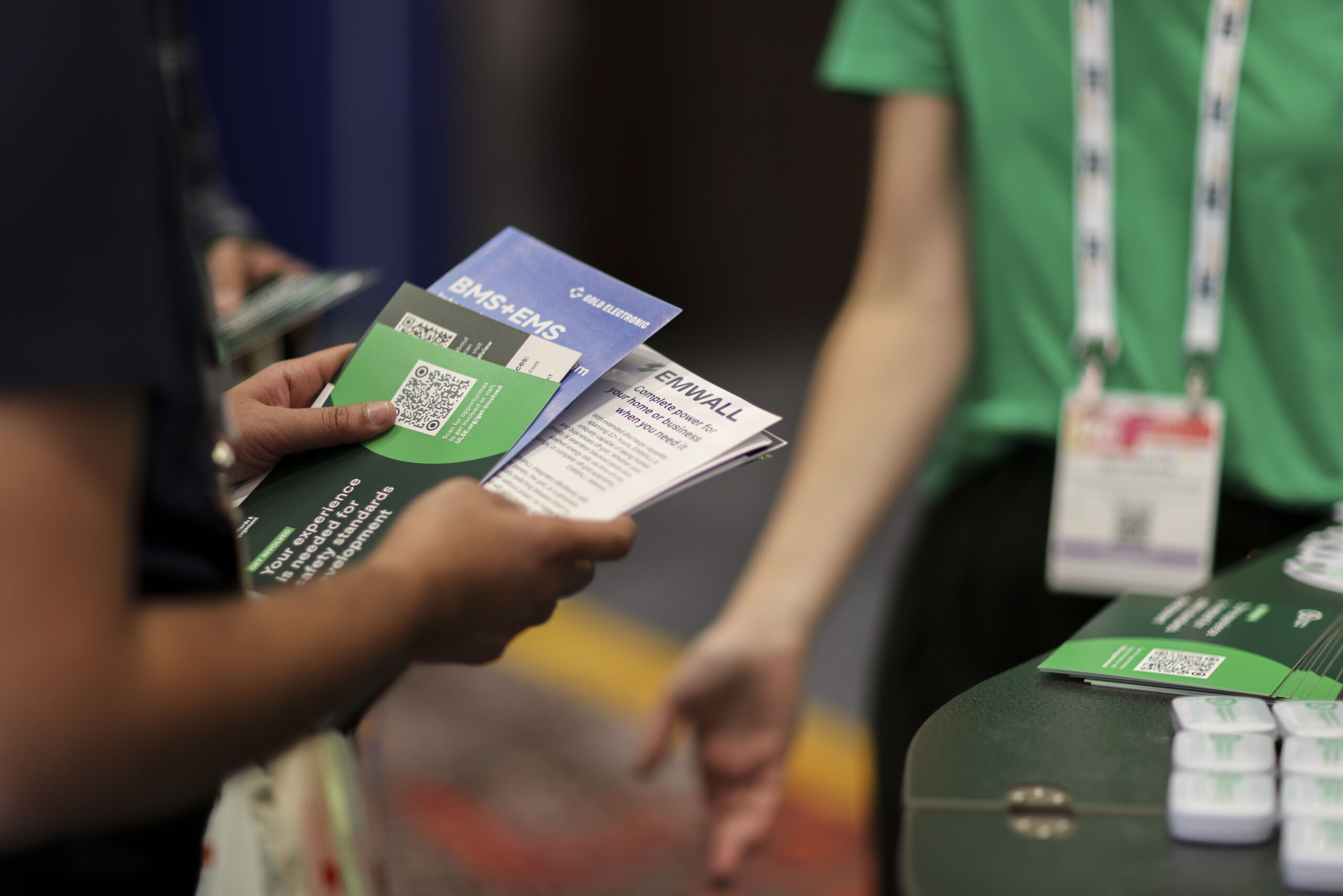 A person holding event brochures with QR codes, speaking to another person wearing a green shirt and a lanyard with a badge at a registration or info desk.