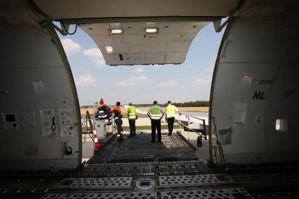 View from inside a large cargo plane with its rear door open, showing five workers in safety vests standing on the loading ramp, looking out toward the runway and sky.