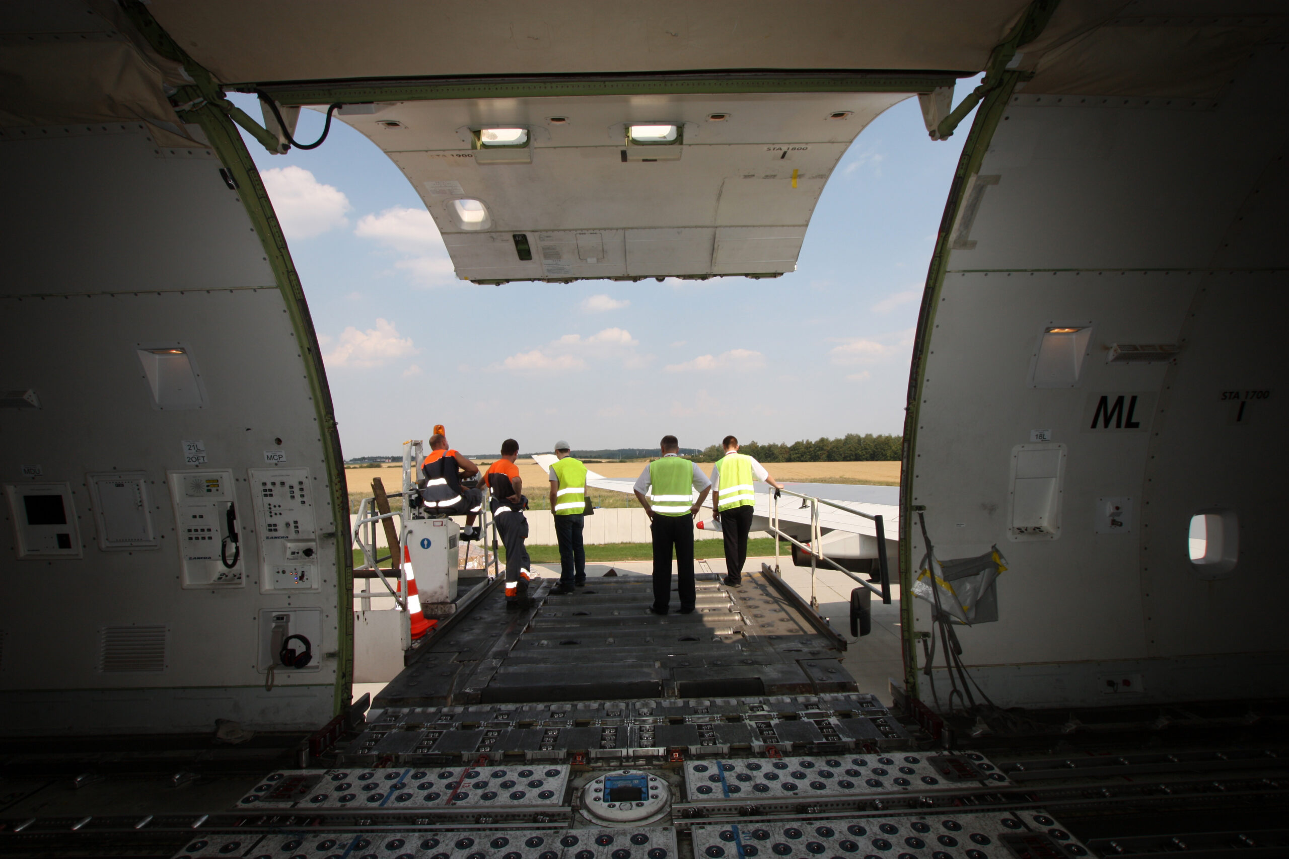 View from inside a large cargo plane with its rear door open, showing five workers in safety vests standing on the loading ramp, looking out toward the runway and sky.
