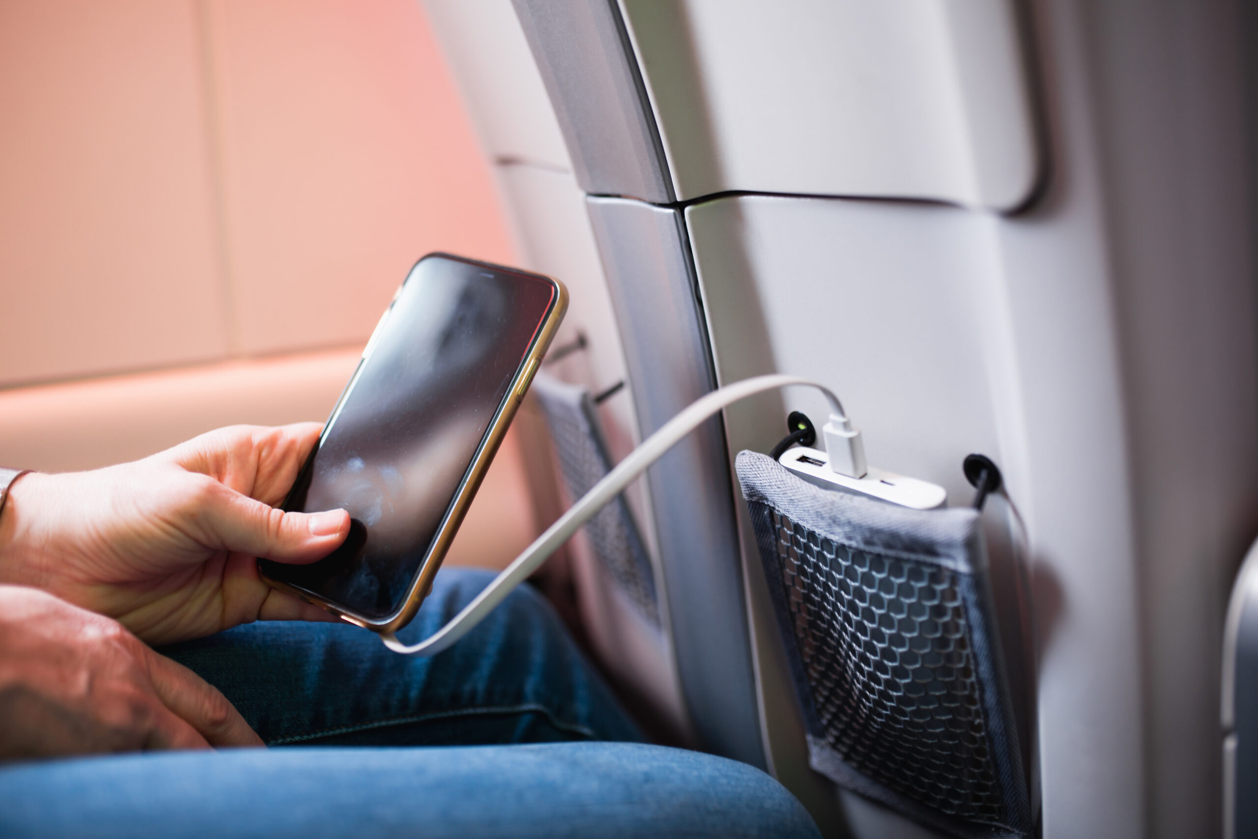 A person sitting on an airplane charges a smartphone using a USB cable plugged into a port on the seatback in front of them. The phone is held in one hand, and a seat pocket with a mesh front is visible.