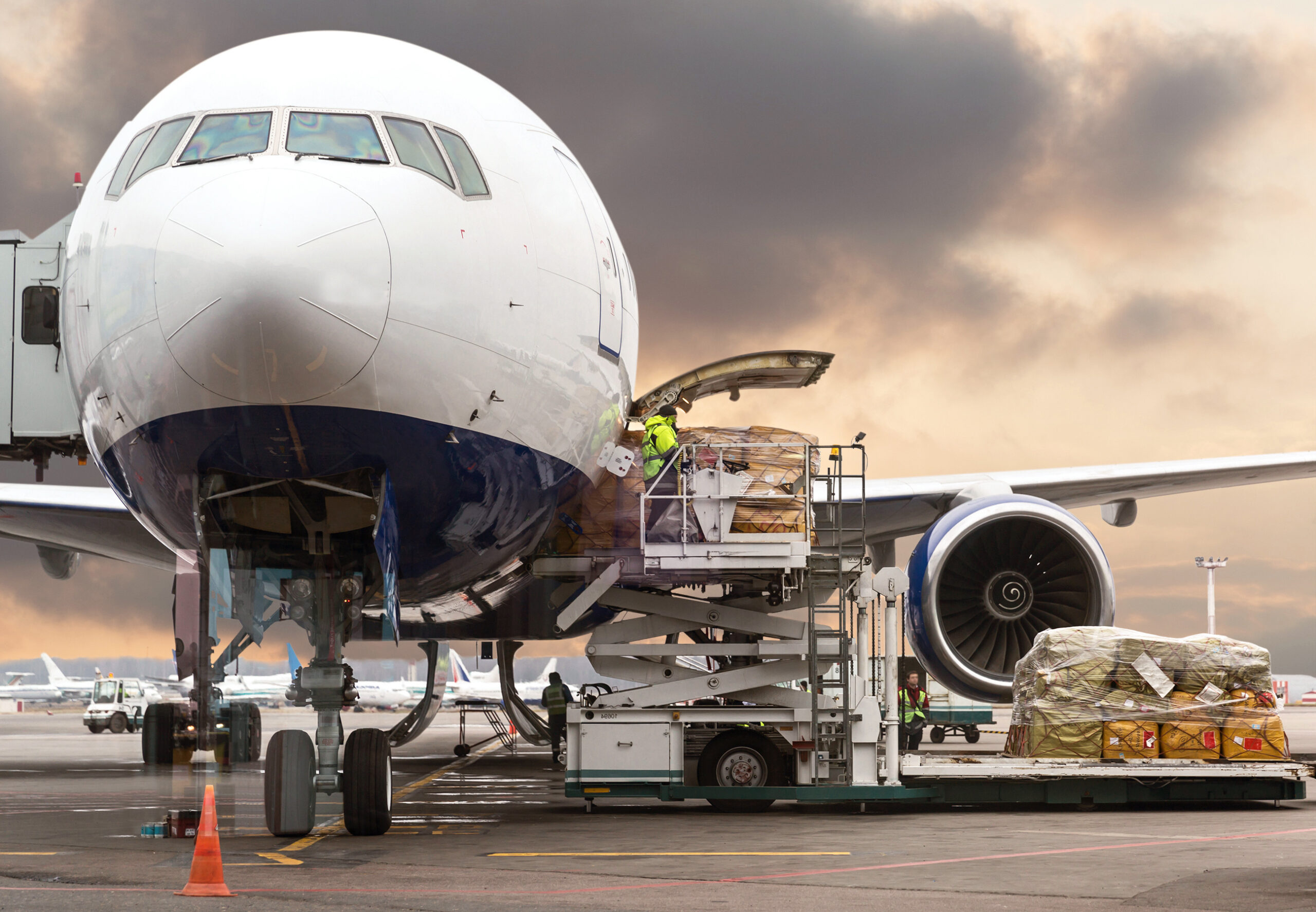 A cargo airplane is being loaded with freight using a conveyor belt vehicle at an airport. A worker in a reflective vest manages the process. The sky is cloudy and other cargo is visible nearby.