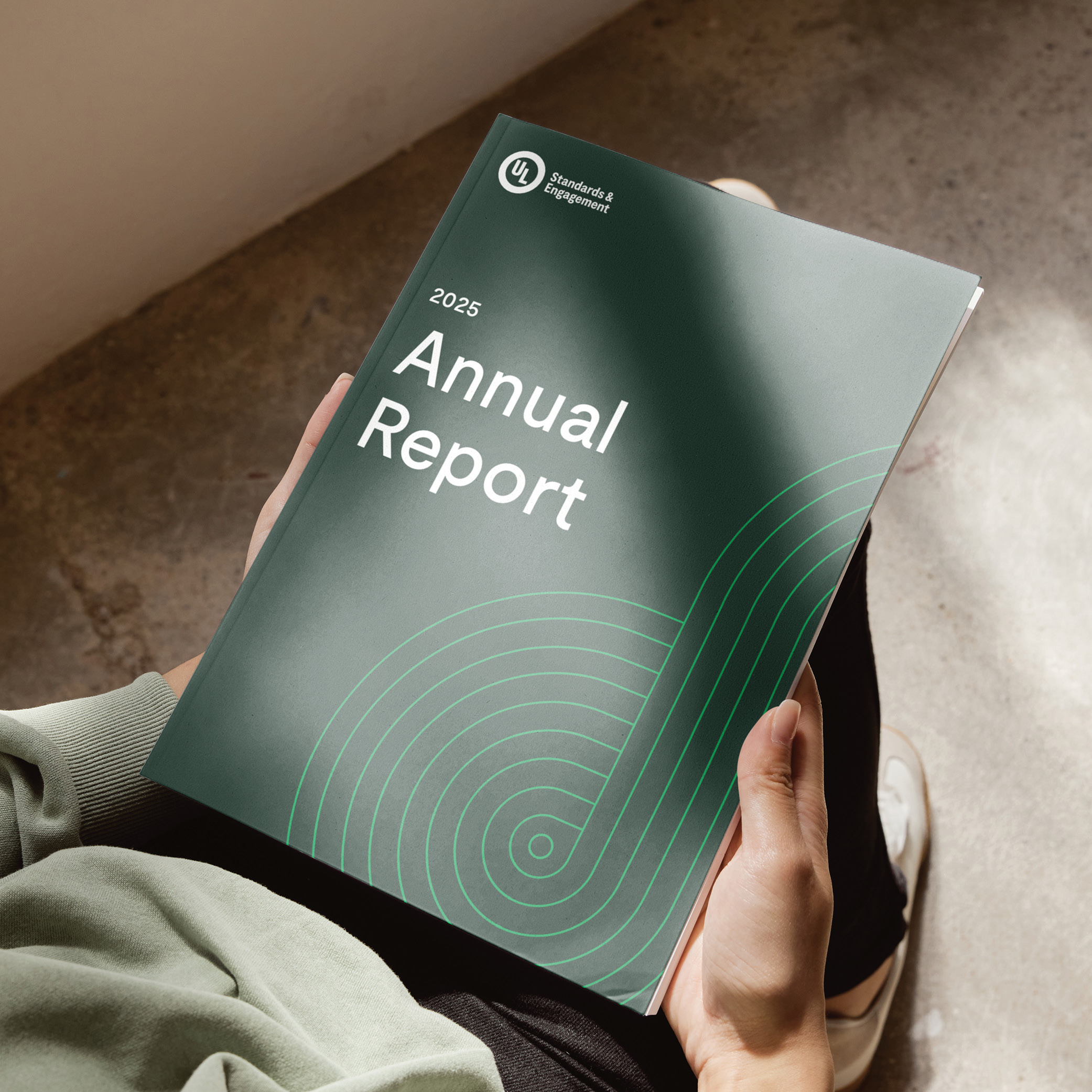 A person holding a green 2025 Annual Report with abstract circular lines on the cover, sitting on a concrete floor with sunlight casting a shadow across the report.
