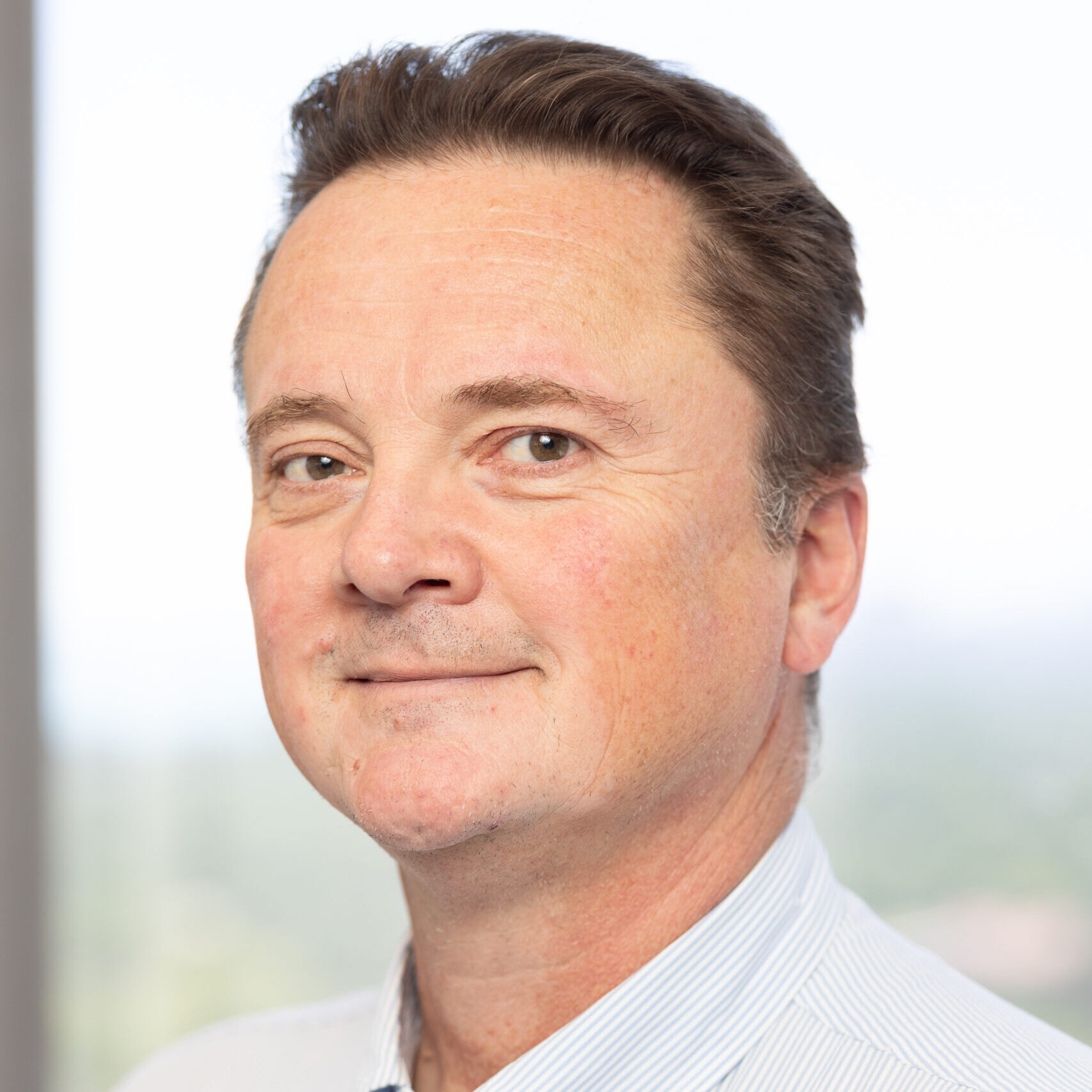 A middle-aged man with short brown hair, wearing a light blue button-up shirt, smiles softly while posing indoors against a blurred, bright background.