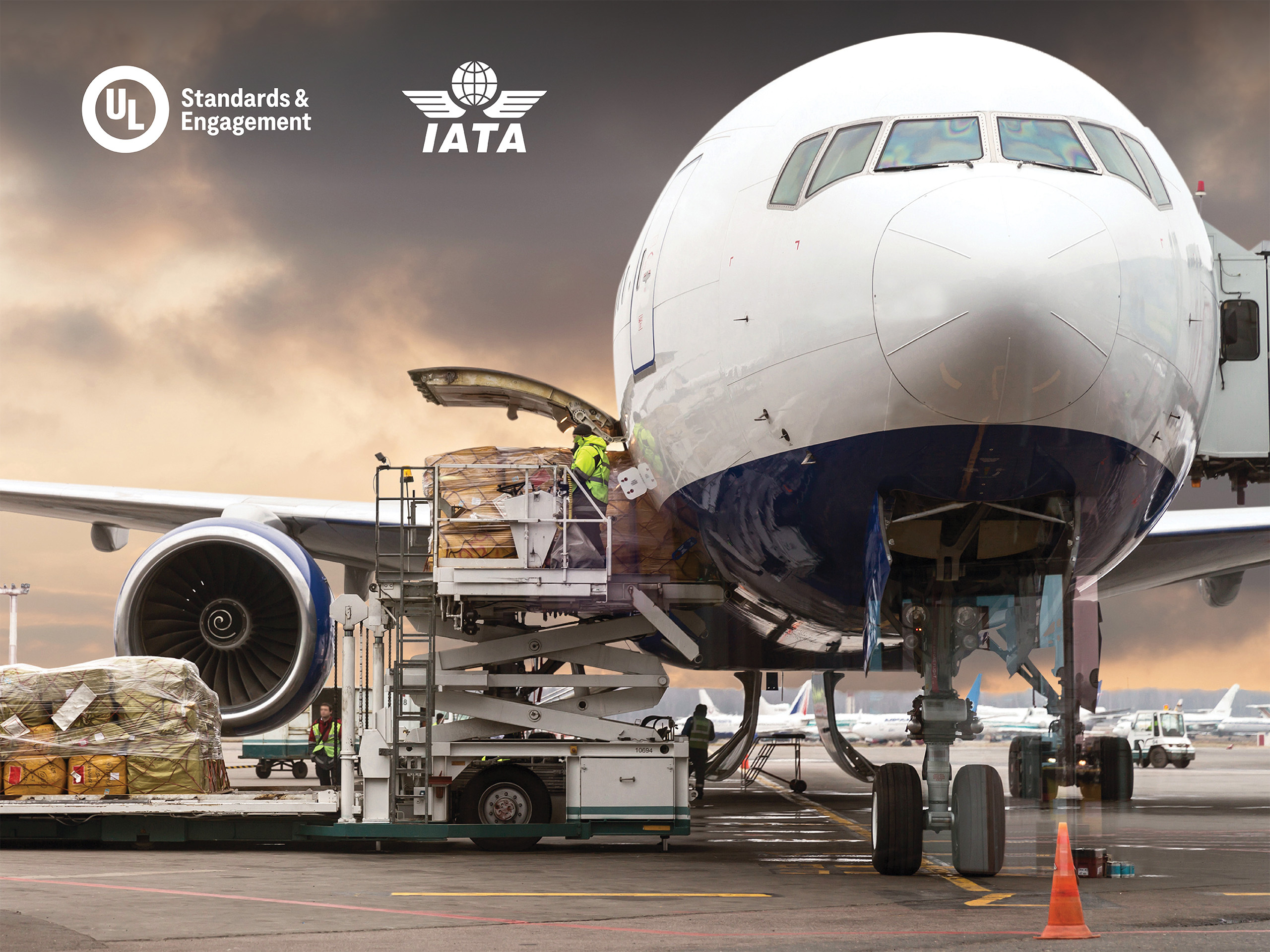 A large cargo airplane is being loaded with freight at an airport. Workers and equipment are visible, with boxes being moved into the plane. The UL and IATA logos are shown in the upper left corner against a cloudy sky.
