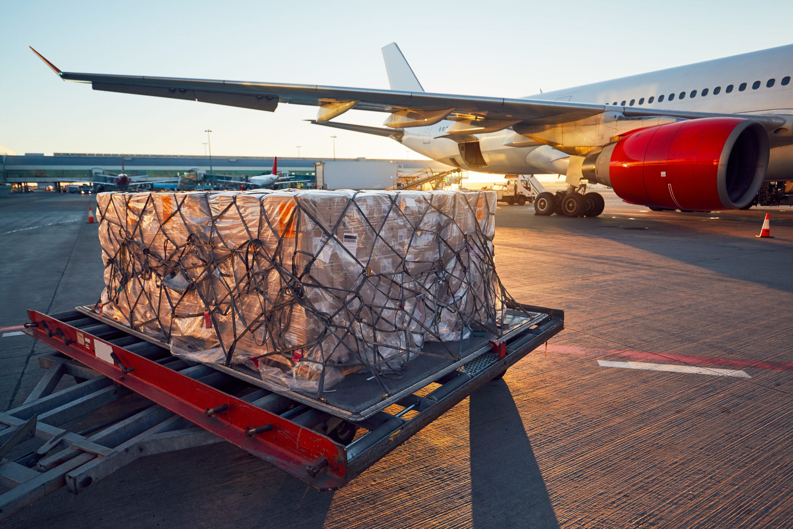 A large pallet of cargo wrapped in plastic and netting sits on a loading platform next to a commercial airplane with red engines at an airport during sunrise.