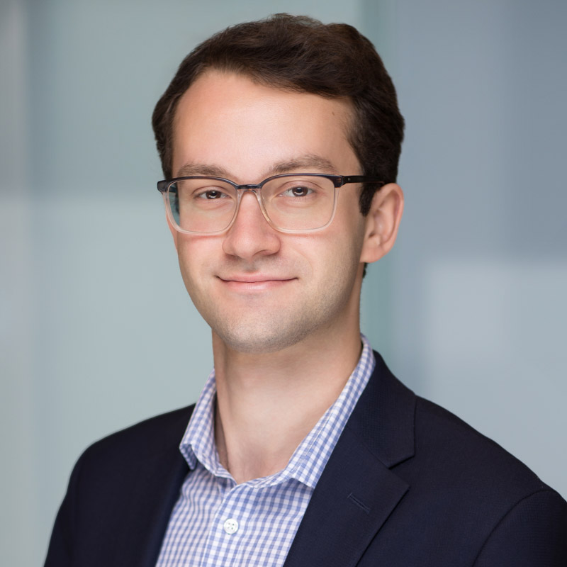 A young man with short brown hair and glasses, wearing a dark blazer over a checked shirt, smiles slightly while posing in front of a blurred light blue background.