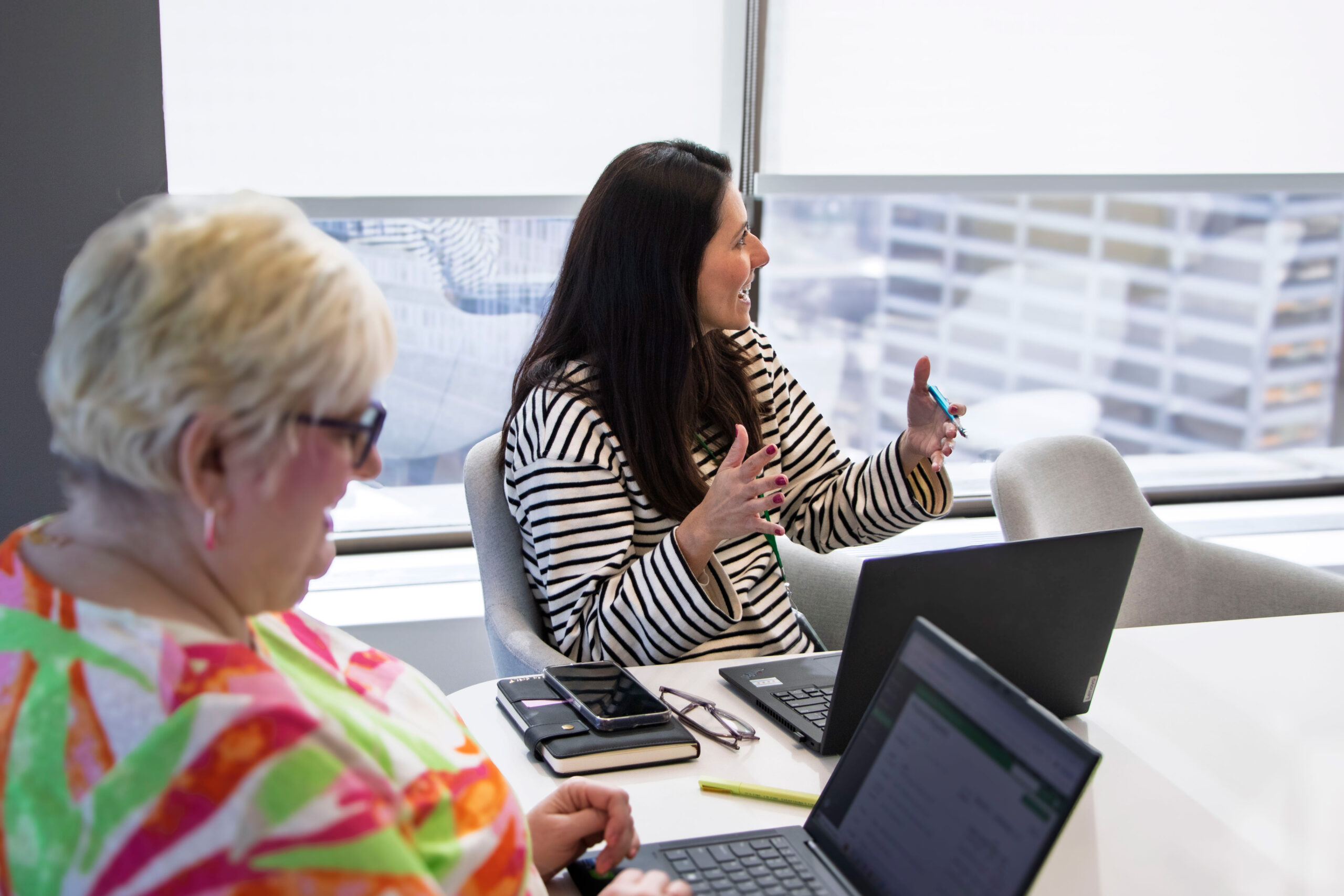 Two women sit at a table with laptops and notebooks, having a discussion in a bright office with large windows. One woman gestures while talking; the other types on her laptop.