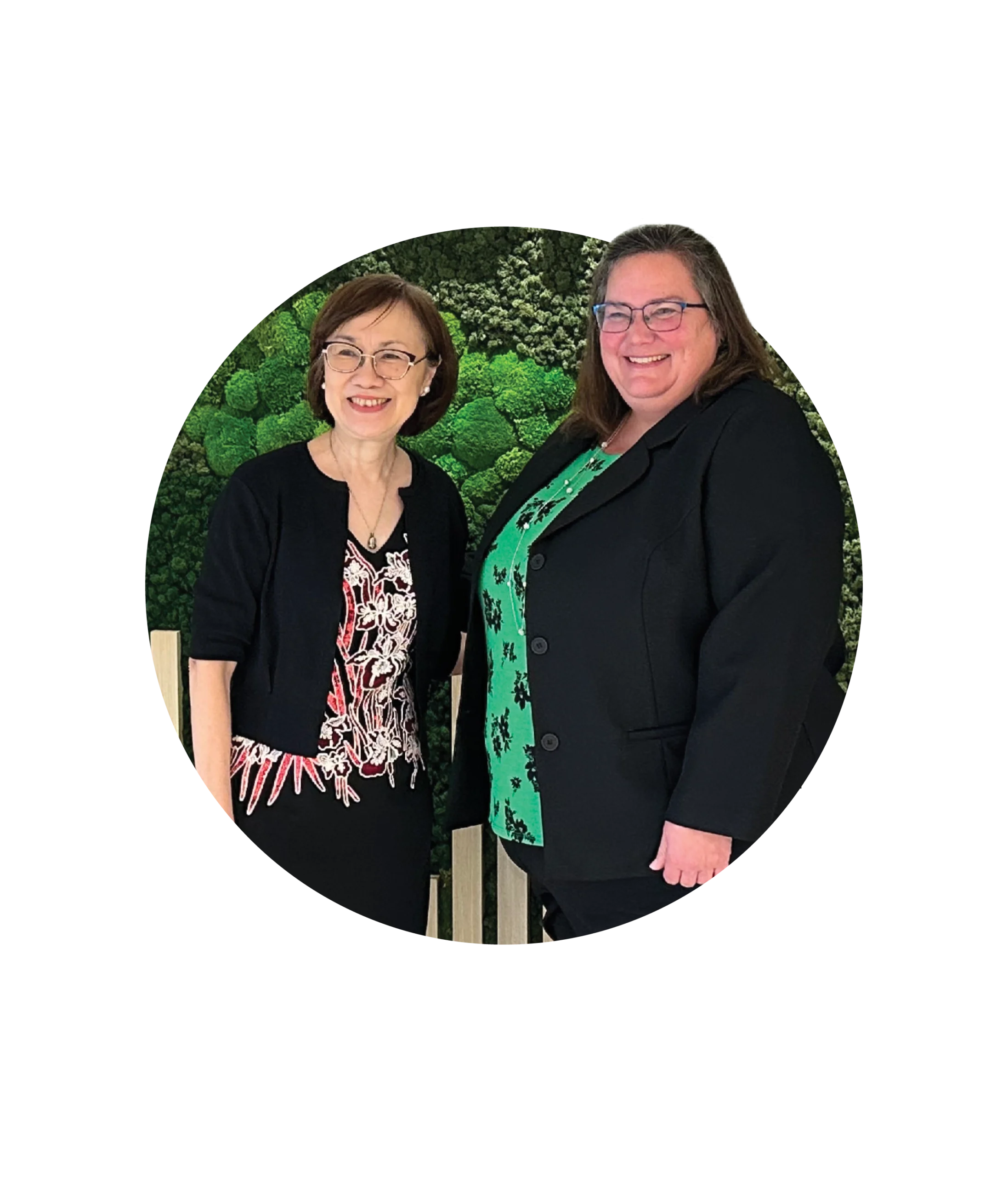 Two women stand side by side, smiling, in front of a green leafy background. Both wear glasses and black blazers—one in a floral dress, the other in a green top with black patterns—reflecting their commitment to global safety partnerships.