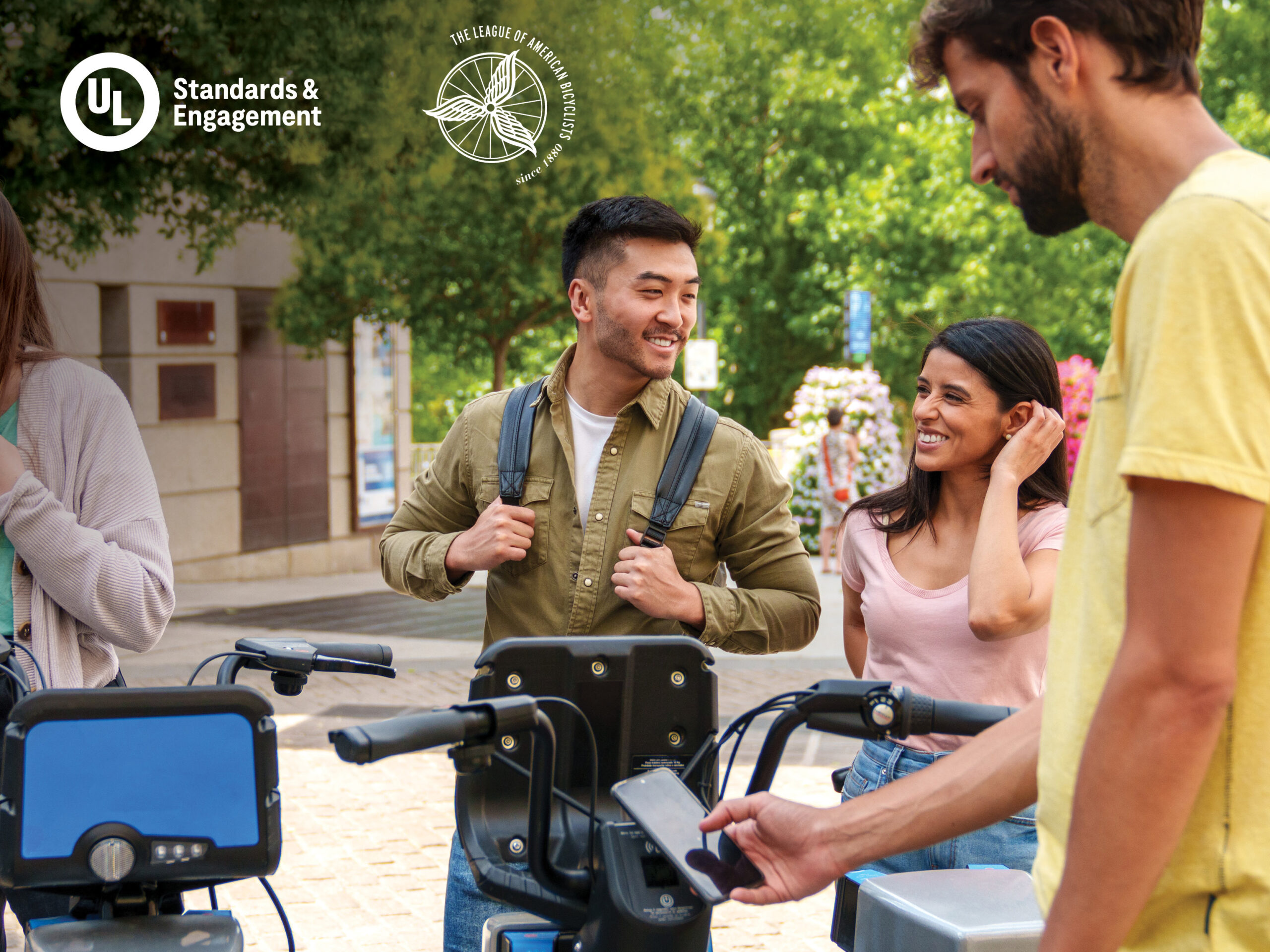Three people stand by electric rental bikes outdoors, smiling and talking. Another person in a yellow shirt uses a smartphone at the bike station. Logos for UL Standards & Engagement and The League of American Bicyclists appear in the corner.