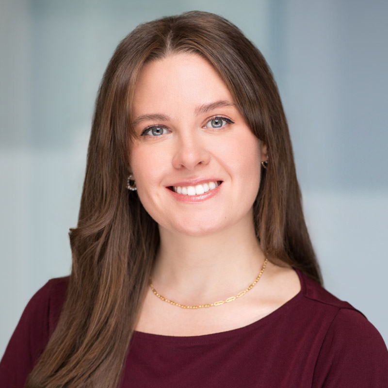 A woman with long brown hair, blue eyes, and a friendly smile wears a maroon top, a gold chain necklace, and small earrings. She is posed in front of a softly blurred, light-colored background.