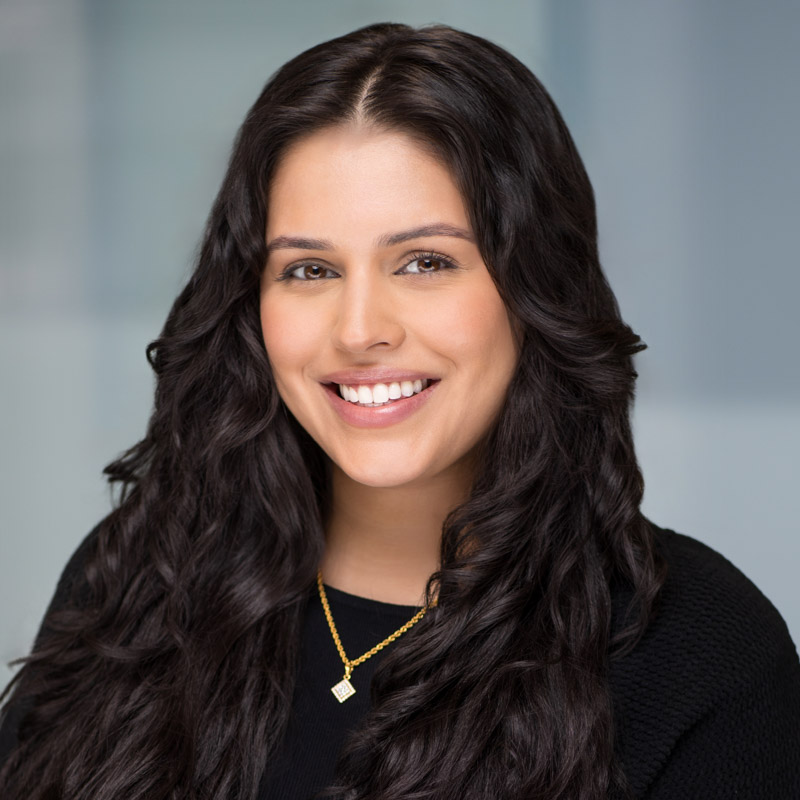 A woman with long, wavy dark hair and a warm smile wears a black top and a gold necklace with a square pendant, posed against a softly blurred, light-colored background.