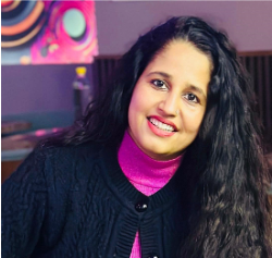 A woman with long, wavy dark hair, wearing a black sweater over a bright pink turtleneck, smiles at the camera. The softly lit background with colorful, abstract wall art reflects her commitment to global safety partnerships.