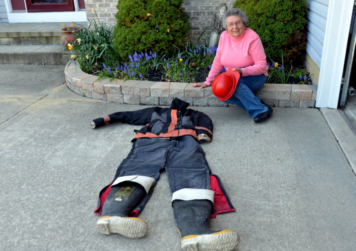 An elderly woman in a pink sweater sits on a curb holding a red firefighter helmet, smiling next to a firefighter suit laid out flat on the driveway. Flowers bloom in the garden behind her.