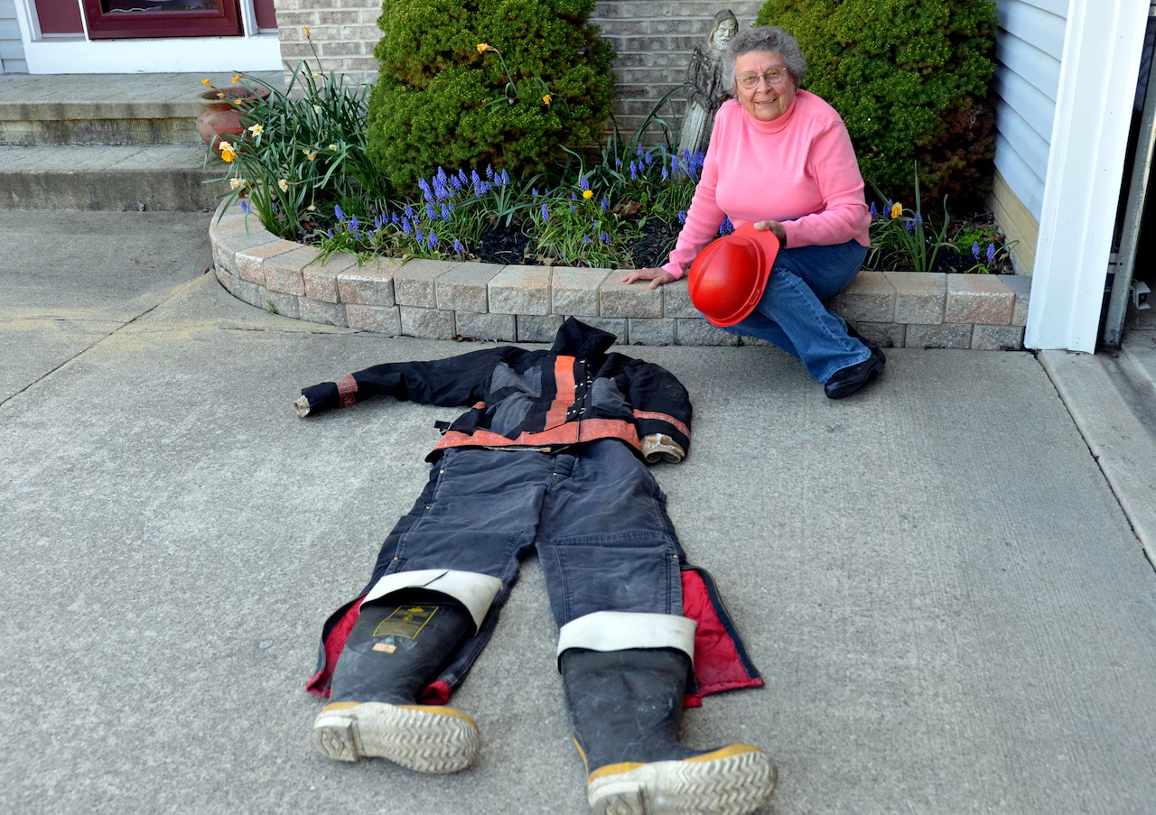 An elderly woman in a pink sweater sits on a curb holding a red firefighter helmet, smiling next to a firefighter suit laid out flat on the driveway. Flowers bloom in the garden behind her.