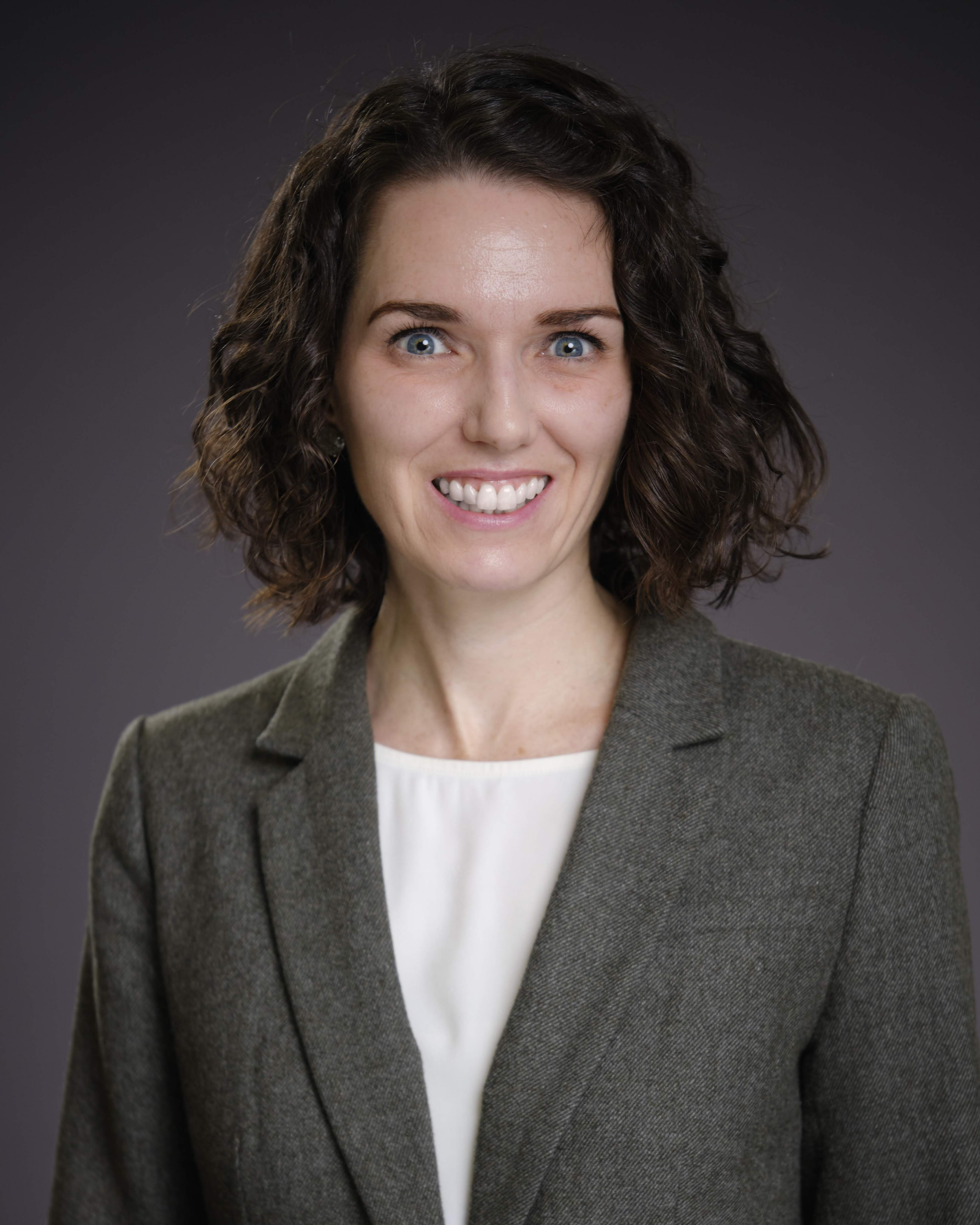 A woman with curly brown hair wearing a grey blazer and white blouse smiles at the camera against a plain, dark background.